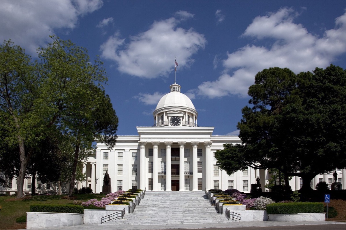 A white domed capitol building standing at the top of a long flight of stone steps, mature trees framing it on both sides and beds of pink flowers along the terrace under a bright sky.