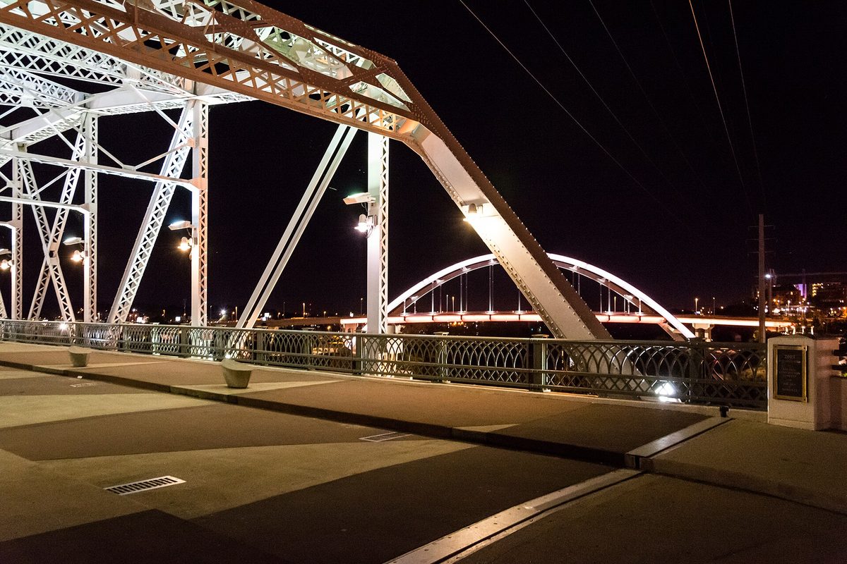 Nashville's pedestrian bridge deck at night, white steel truss and ornate railing lit up, with an illuminated arch bridge beyond.