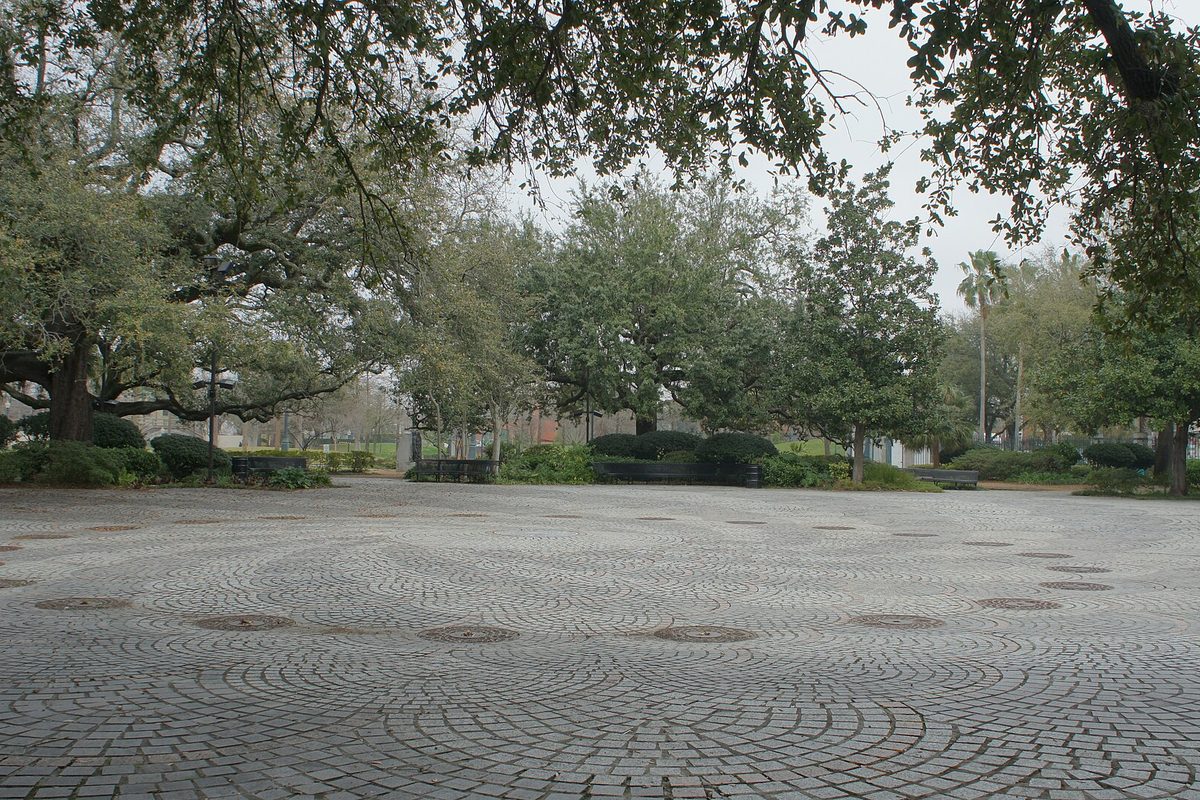 New Orleans park plaza paved in swirling brick circles, ringed by live oaks, clipped hedges and benches under an overcast sky.