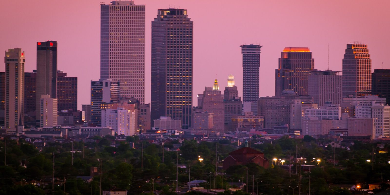 New Orleans downtown towers against a pink twilight sky, lit windows above a low green tree canopy and neighborhood rooftops