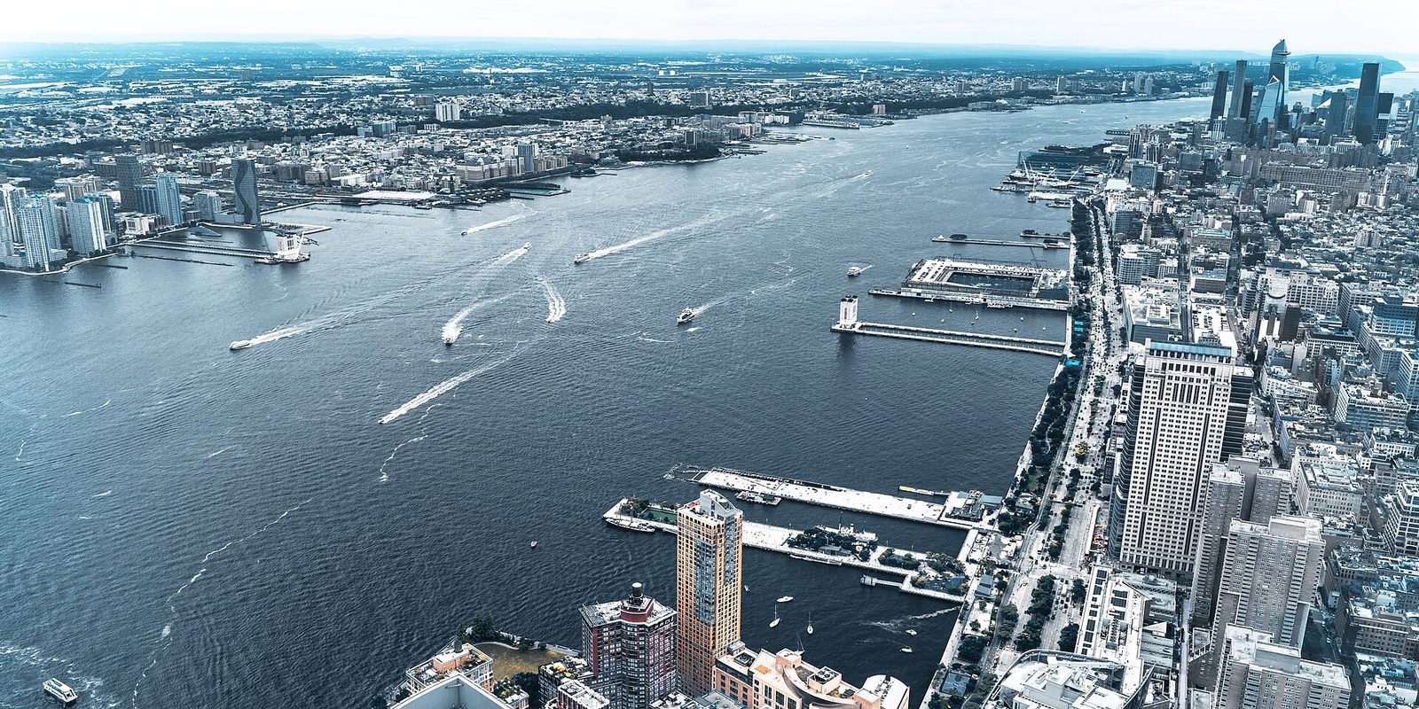 New York from high above the Hudson River, Manhattan piers and towers along the right bank, New Jersey opposite, boat wakes crossing the water