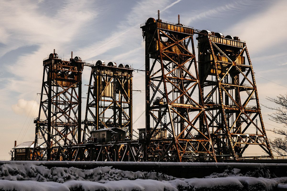 Newark's rusted steel vertical-lift railroad bridge towers standing over snow-covered tracks, catching low sun under a contrail-streaked sky.