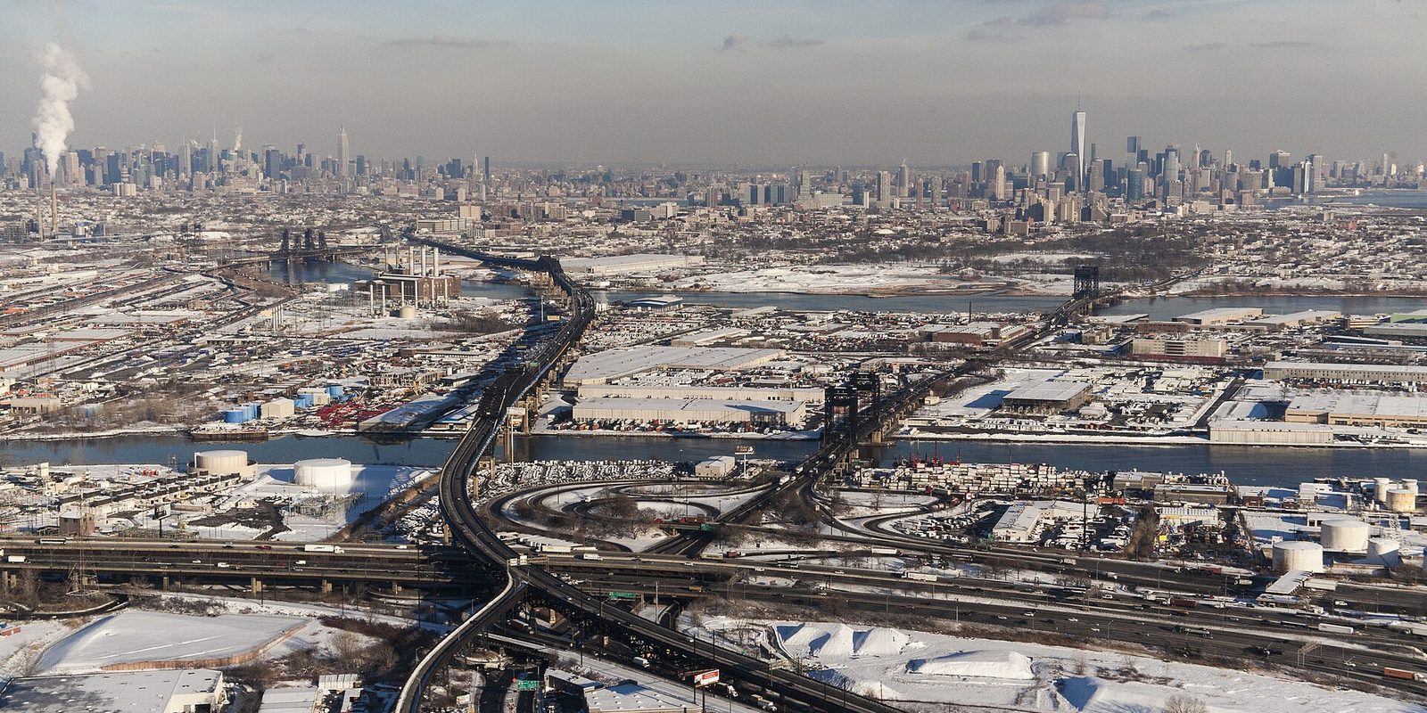 Newark from the air in winter, snow over port yards, a river and a highway interchange, with the hazy Manhattan skyline far beyond