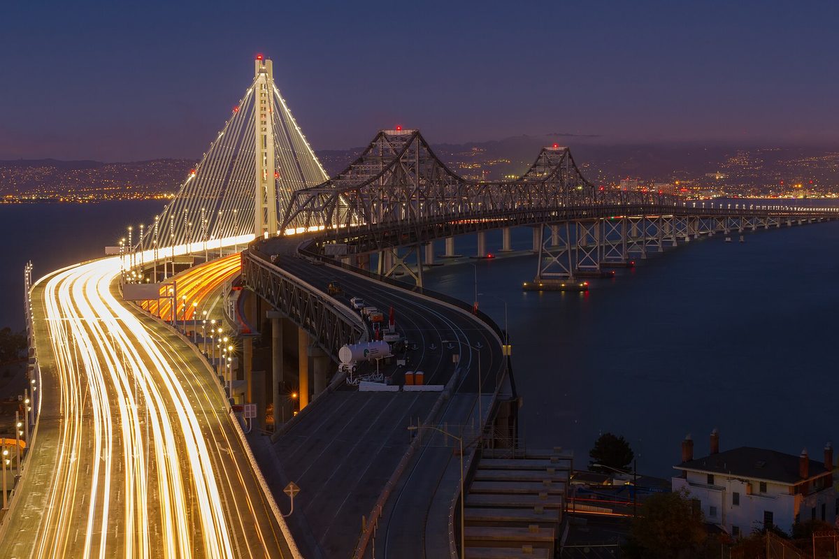 The Bay Bridge at dusk from Oakland, its lit suspension tower and older cantilever span beyond, headlight trails on the approach