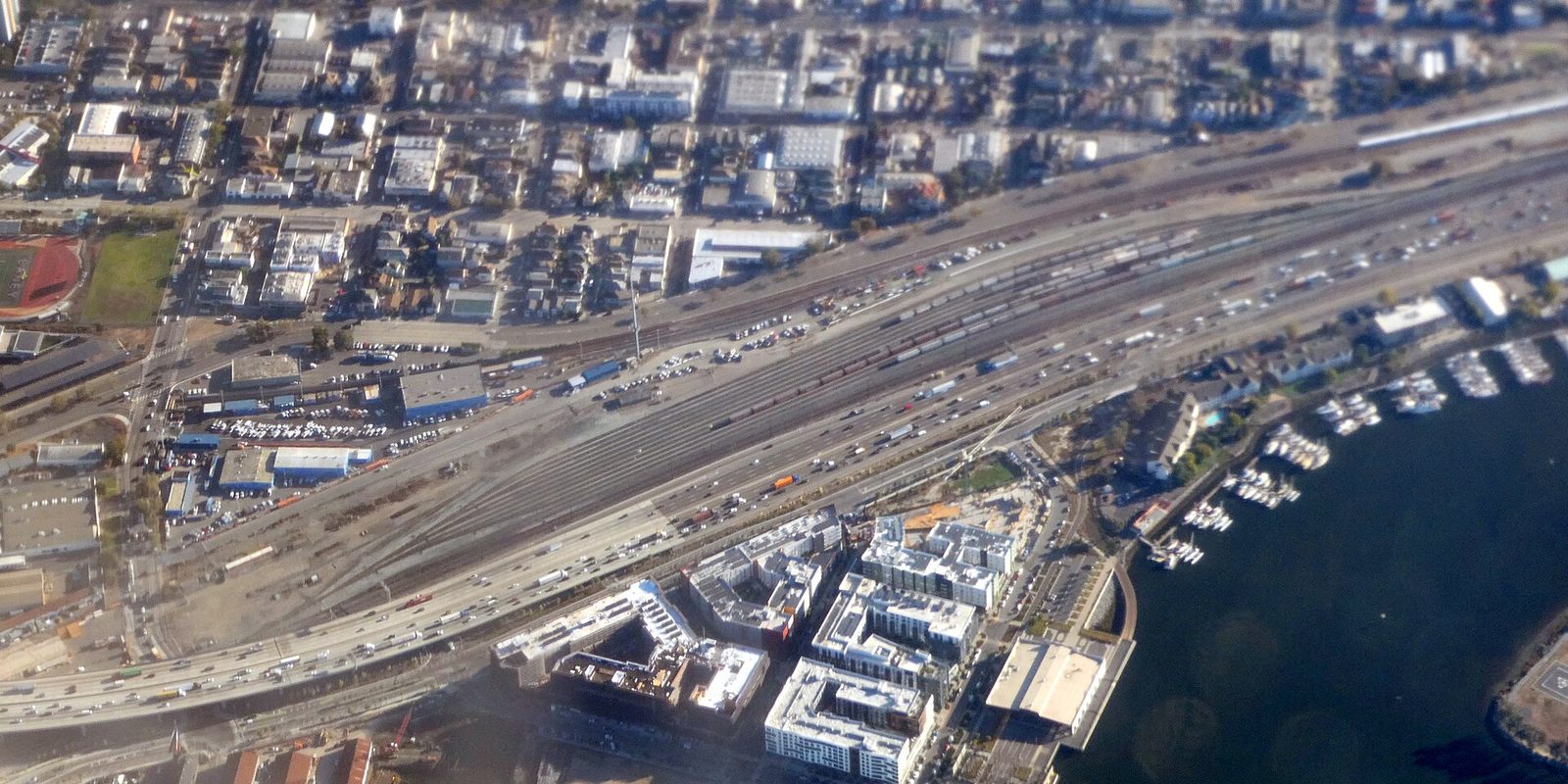 Oakland from the air, a wide rail yard and freeway running past industrial blocks to a marina of white boats on the waterfront in bright sun