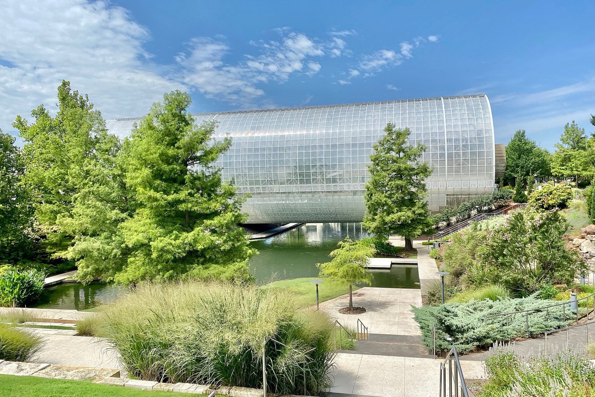 A long curved glass conservatory in the Oklahoma City botanical gardens seen across a still lake, with conifers, ornamental grasses and paved terraces in front of it.