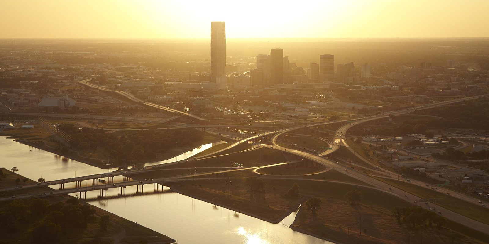 Oklahoma City backlit by a low sun, downtown towers hazy on the horizon above a sweeping highway interchange and a river crossed by low bridges