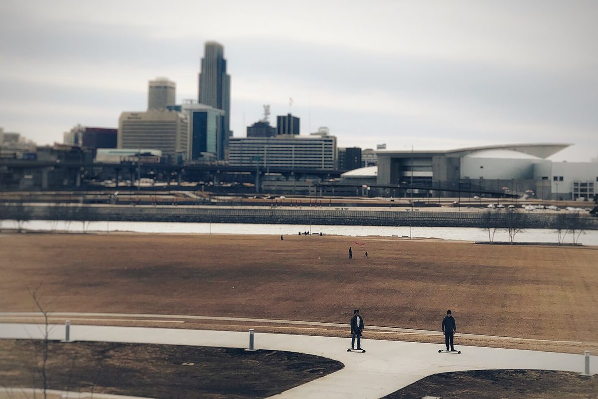 Two skateboarders roll along a paved path beside Omaha's dormant riverfront lawn, the Missouri River and downtown towers under a flat grey sky.