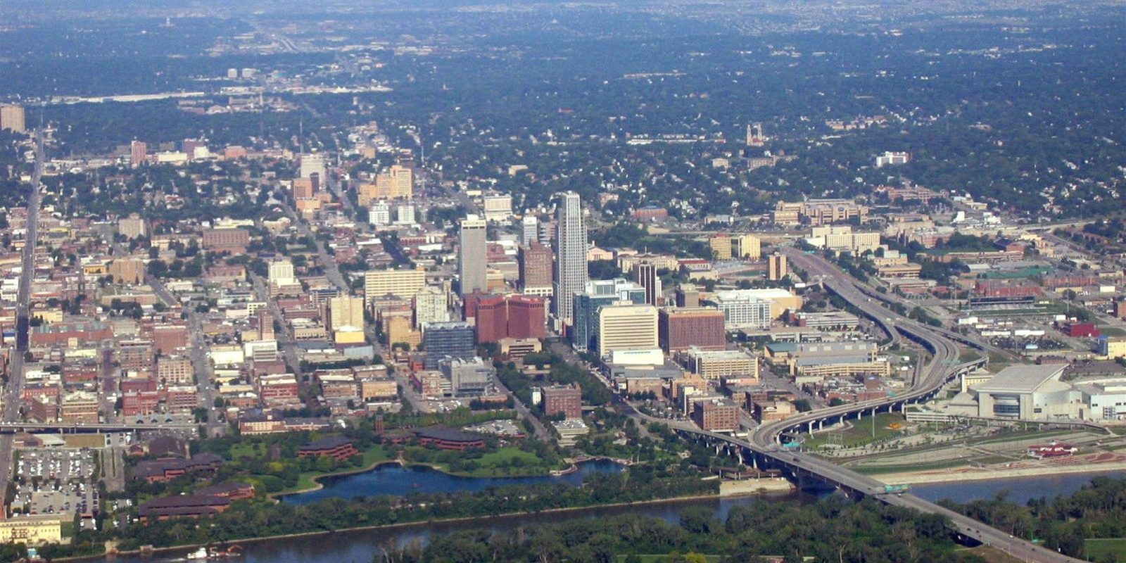 Omaha from the air, downtown blocks and a few towers surrounded by green summer tree canopy, an elevated highway and a bridge over the river