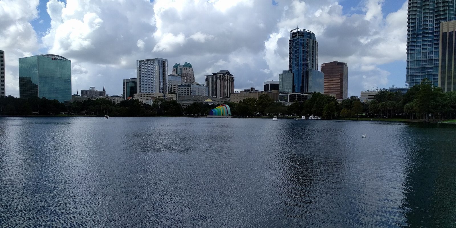 Orlando's downtown skyline across a lakefront park, with a rainbow-striped bandshell and swan boats on the far shore under tall white clouds