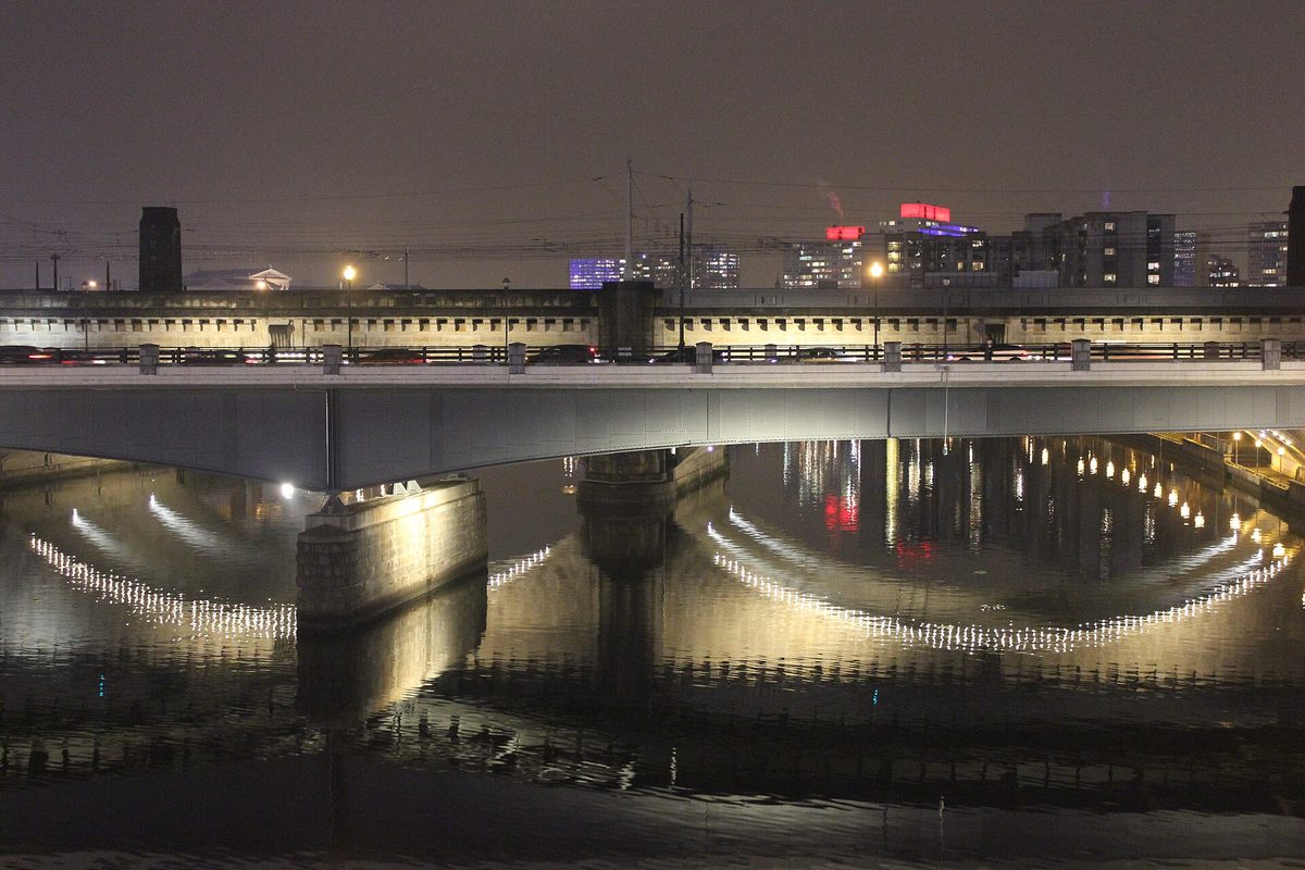 Philadelphia at night: a lit road bridge and rail viaduct cross the river, lamps and skyline lights rippling on the black water.