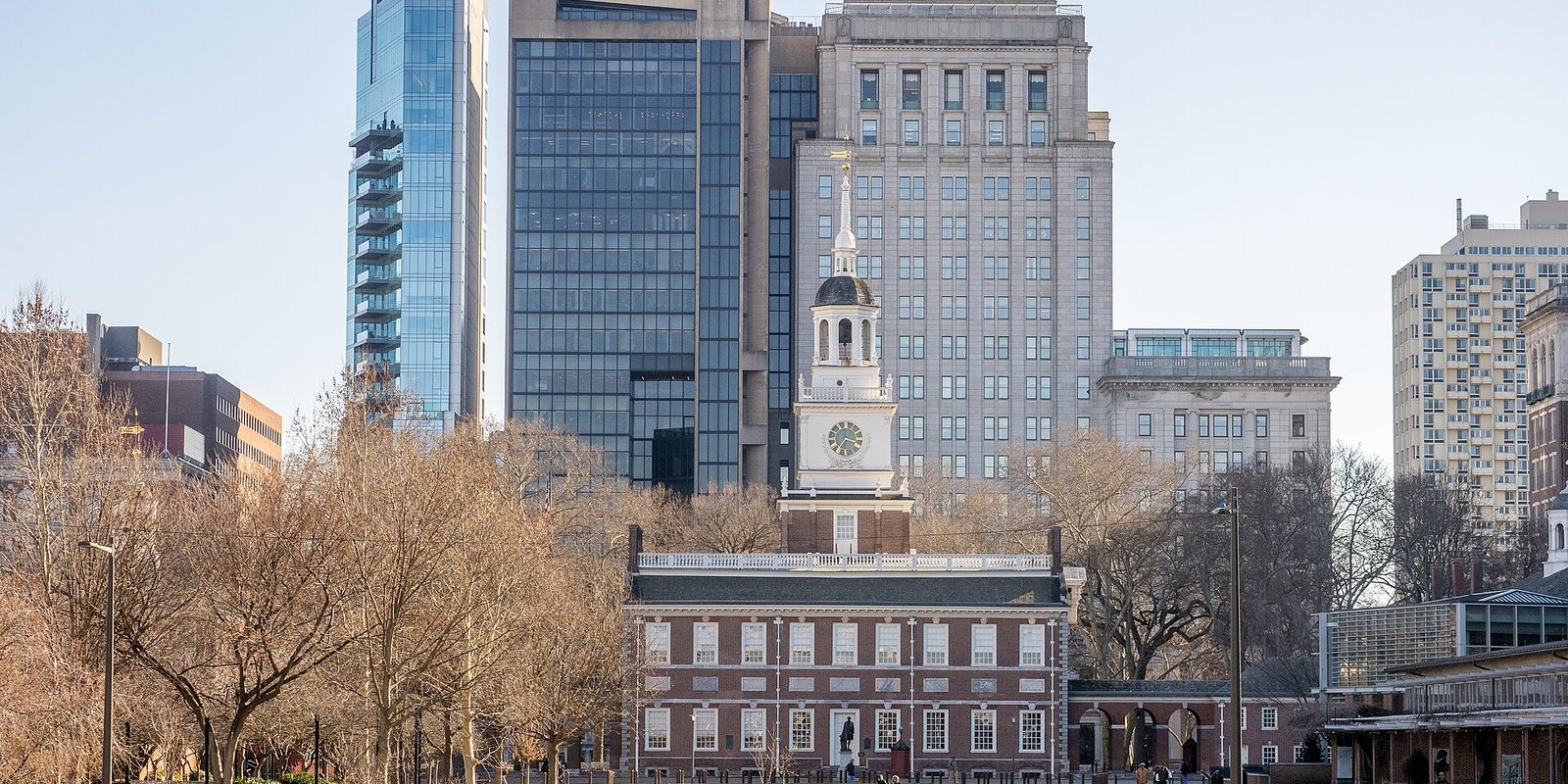 Philadelphia's Independence Hall and its white clock tower behind leafless trees, with glass and stone high-rises close behind under a pale clear sky