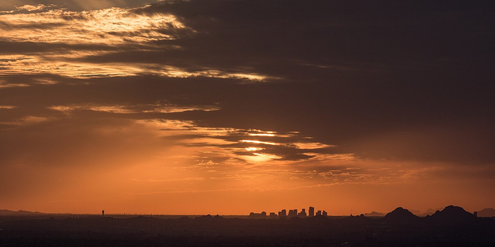 Phoenix's skyline a low silhouette on the valley horizon beside desert buttes, under a burning orange sunset sky streaked with dark cloud