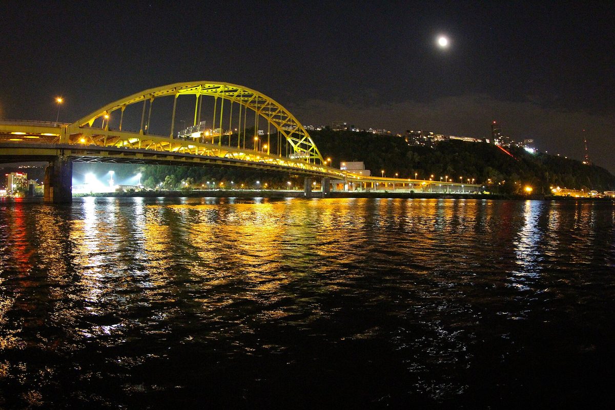 Pittsburgh's yellow steel arch bridge glows over the river at night, a full moon above the wooded hillside and lights rippling on the water.