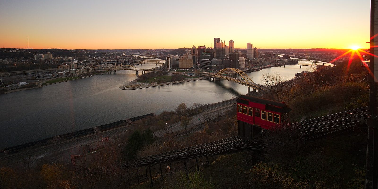 Pittsburgh's downtown at the meeting of the rivers, seen from the hillside past a red incline car on its track, with a low sun flaring at the horizon