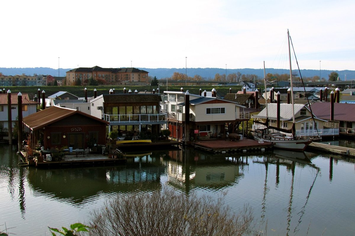 Floating homes and a moored sailboat line a calm Portland marina, with apartment buildings and low wooded hills across the water.