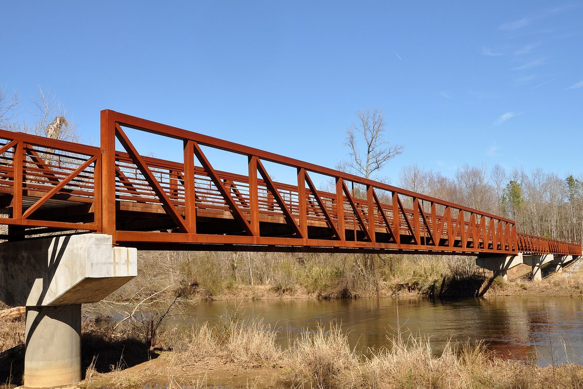 Raleigh, North Carolina: a rust-red steel truss footbridge on concrete piers crossing a slow brown river, with bare trees and dry grass along the banks.