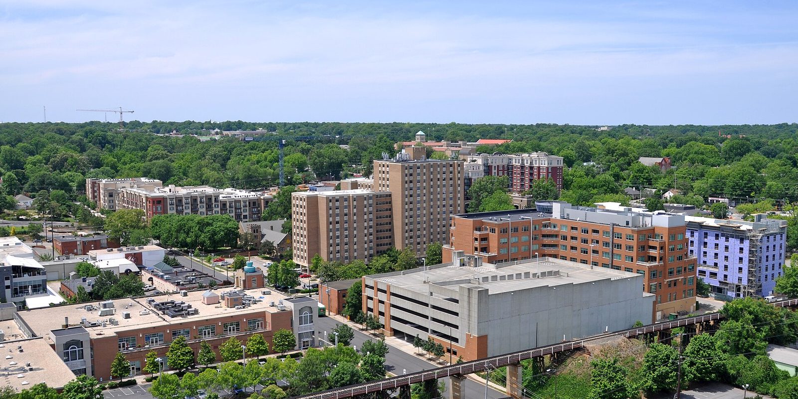 Raleigh from above: mid-rise brick apartment blocks and a concrete parking deck set in dense green summer tree canopy, a construction crane on the horizon