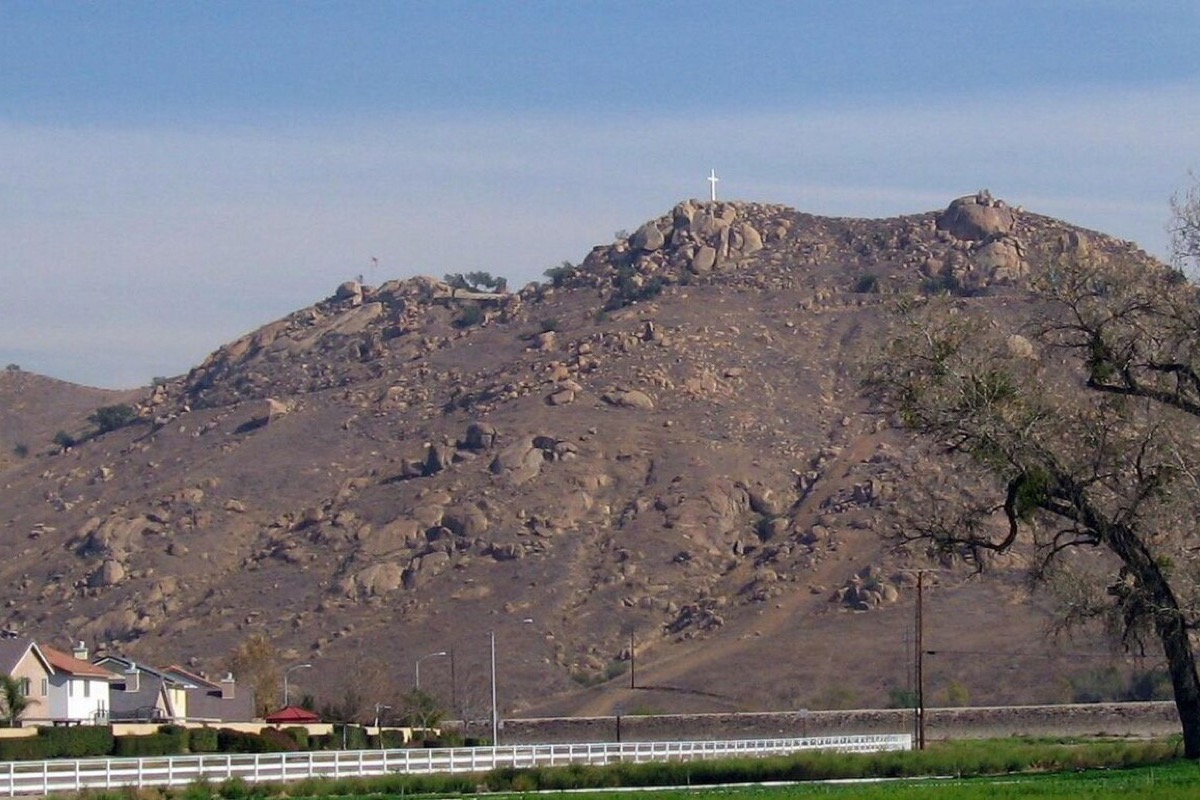 A bare rocky hill rising straight out of the town at Riverside, boulders and dry scrub over its whole face with a cross on the summit, and white fenced houses and an oak along the road below.