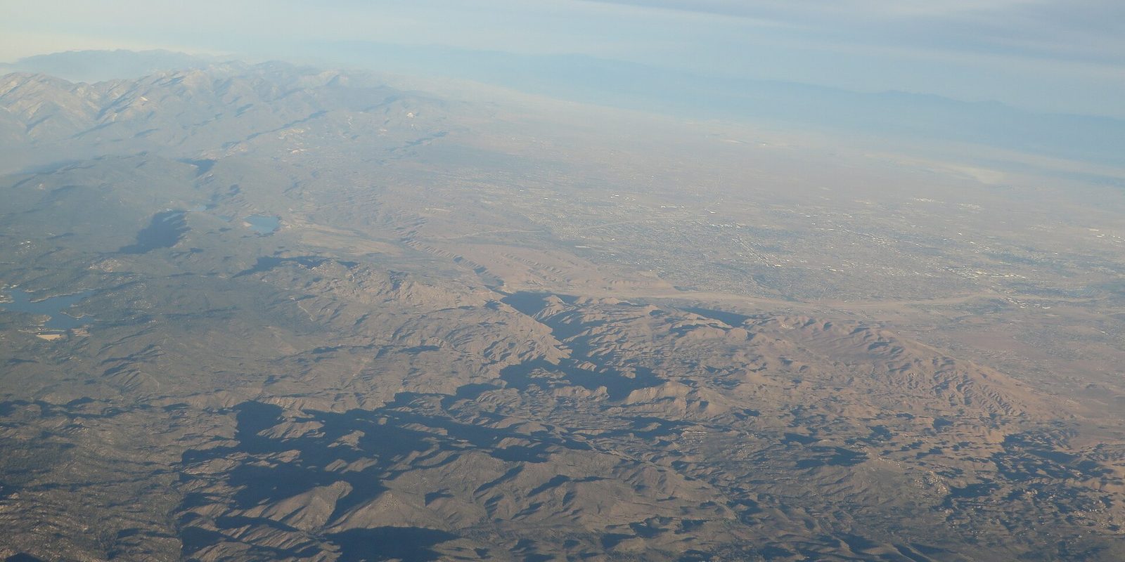 Riverside seen from an aircraft window: hazy brown ridges and a small reservoir in front of the valley's sprawl, mountains fading into the distance