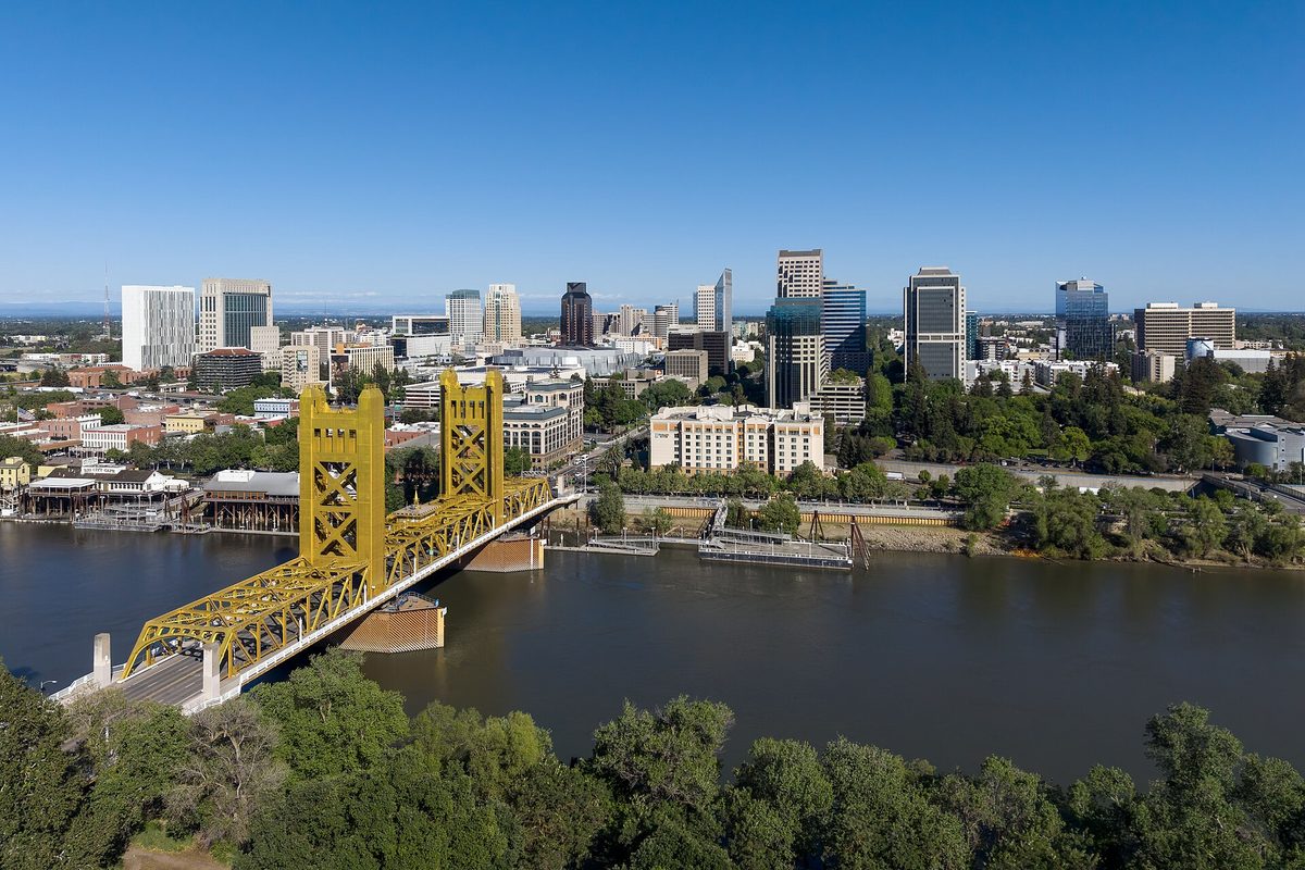 Sacramento, California: the golden Tower Bridge spanning a wide river in front of the downtown high-rise skyline, with leafy treetops in the foreground.