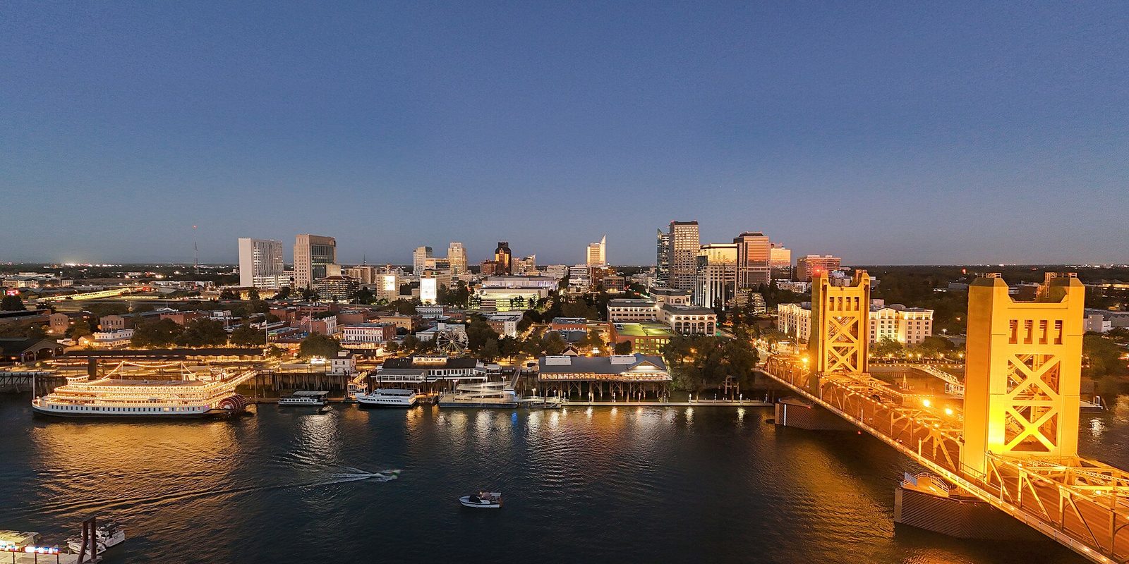 Sacramento at dusk: the lit downtown skyline across the river, a moored paddlewheel riverboat at the old waterfront and the gold lift bridge glowing at right
