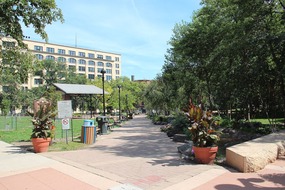 Saint Paul, Minnesota: a brick promenade through a downtown park, with terracotta planters, a rock-lined stream and a cream brick warehouse behind the trees.