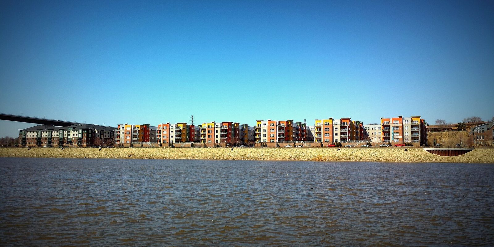 Saint Paul across a wide brown river: a long row of orange, yellow and red mid-rise apartments on a rocky bank under a clear deep-blue sky