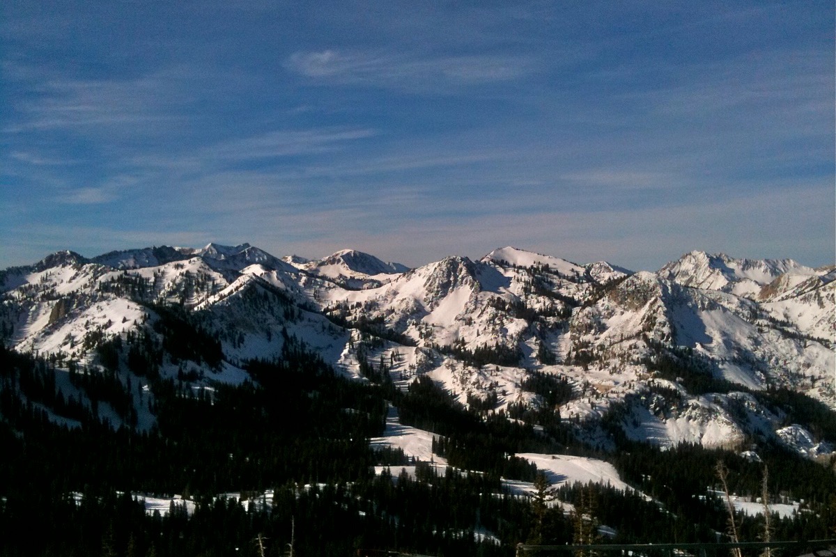 A range of bare granite Utah peaks still holding heavy snow, rising above dark conifer forest under a clear sky.