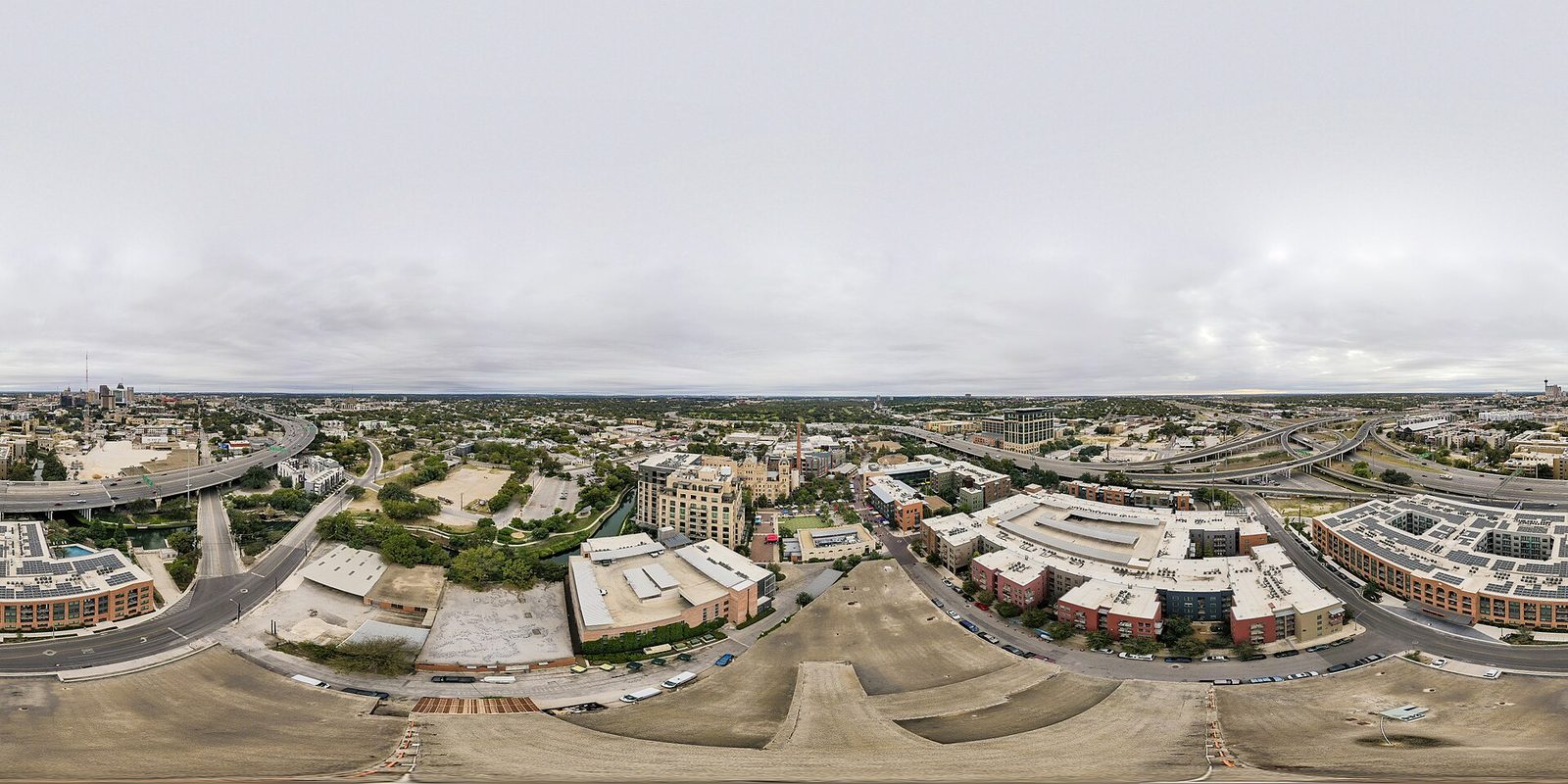 San Antonio from a rooftop under flat grey overcast: sweeping freeway interchanges, low apartment blocks and the river channel, downtown towers far off at left