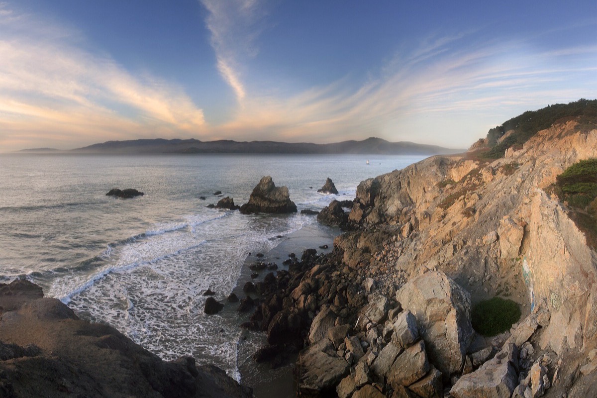 The rocky San Francisco shoreline at the end of the day, surf breaking white around dark boulders, a headland to the right and high cirrus lit gold above the ocean.