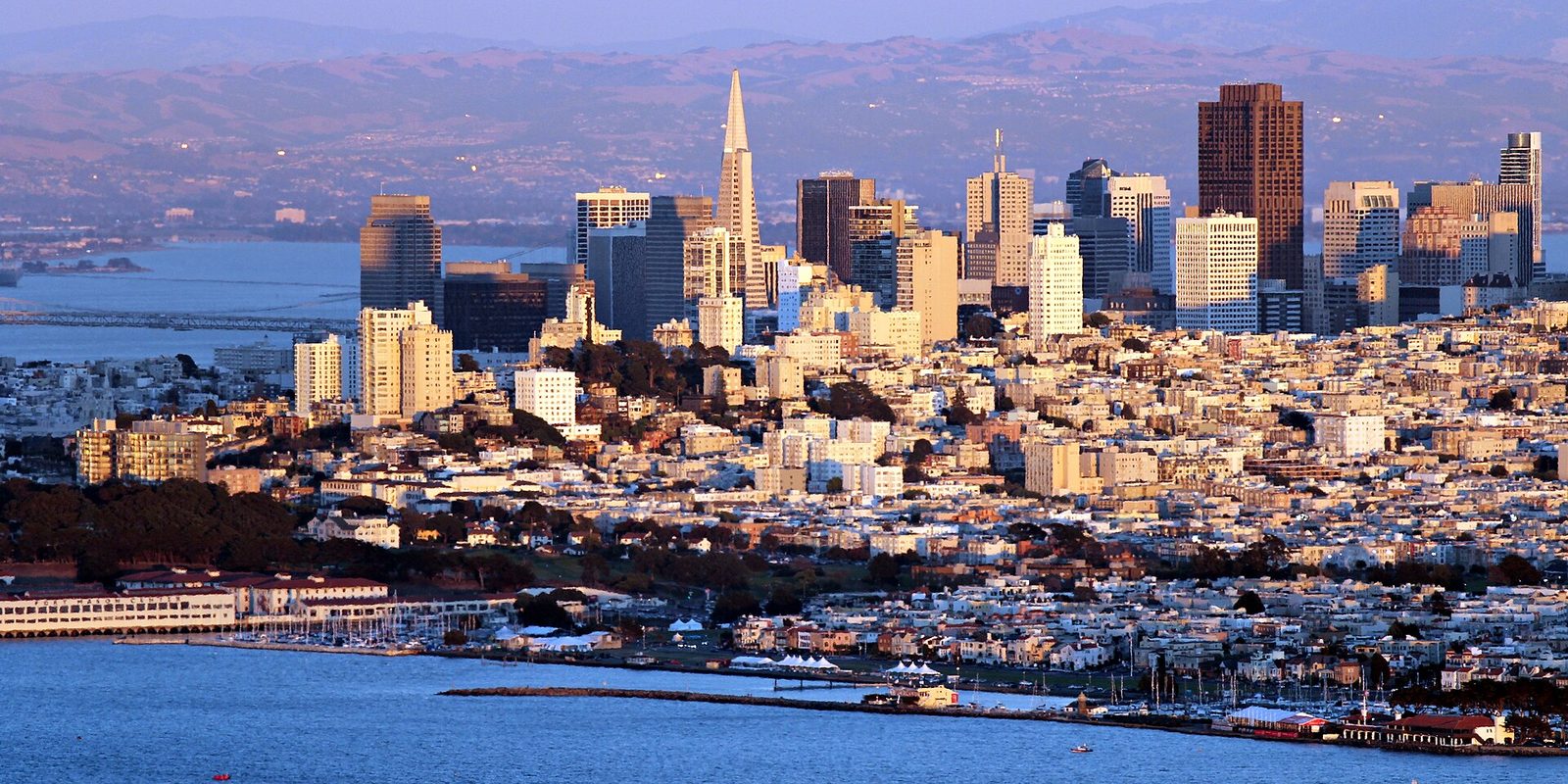 San Francisco from above the bay, dense hillside blocks climbing to downtown high-rises lit by warm low sun, coastal hills behind