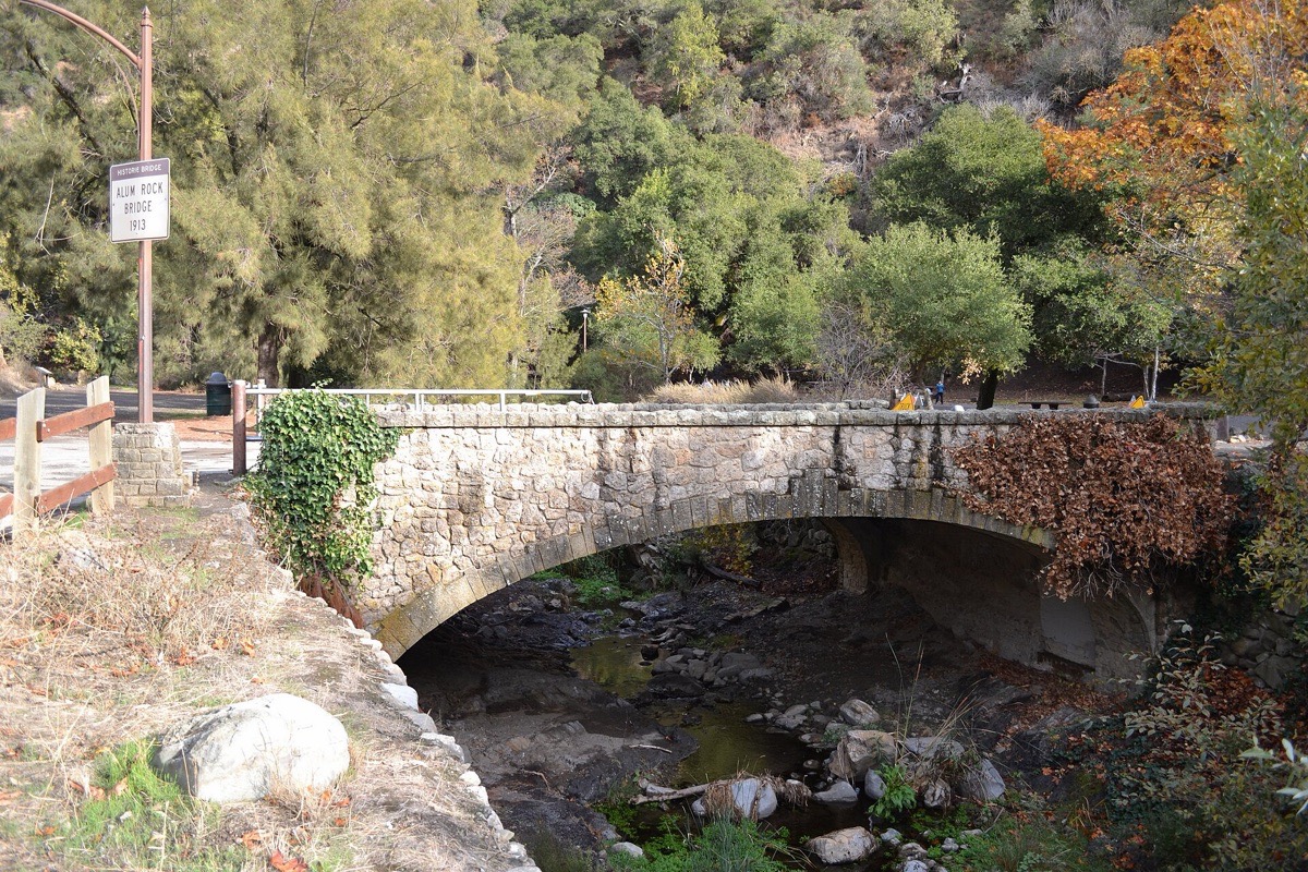 A stone arch bridge crossing a creek in a wooded canyon above San Jose, autumn colour in the trees behind it and boulders in the stream bed below.