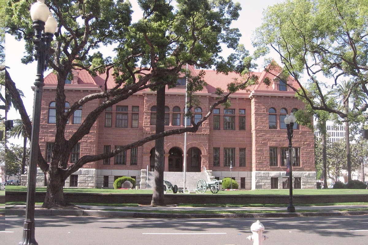 The old red sandstone courthouse at Santa Ana seen across a lawn through the limbs of a large shade tree, its steep red roofs and arched windows catching bright sun.