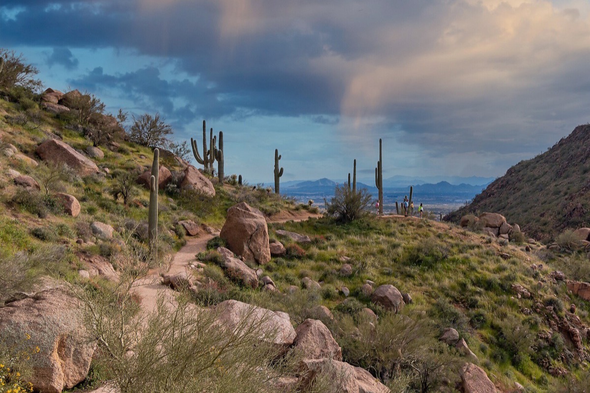 A desert trail climbing between granite boulders and saguaro on the slope below a rock peak north of Scottsdale, with the valley and distant ranges under a heavy sky beyond.