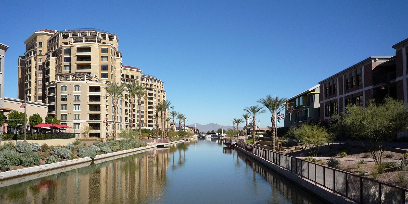 Scottsdale's downtown canal between palm-lined walkways and tan low-rise buildings, desert mountains beyond under a deep blue sky