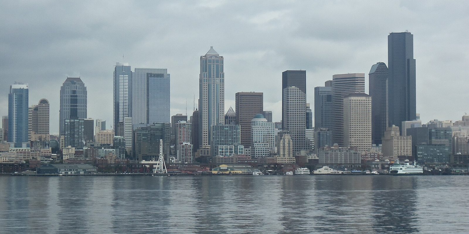 Seattle's downtown skyline seen across the water under heavy grey overcast, with waterfront piers and a docked ferry along the shoreline