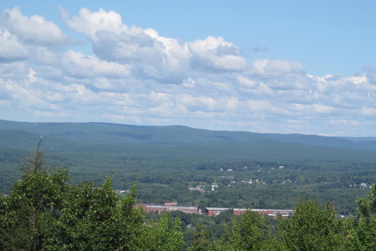 A wide view from a Massachusetts ridge out over forest and a scatter of low buildings to a long blue range of hills under a sky of broken white cloud.