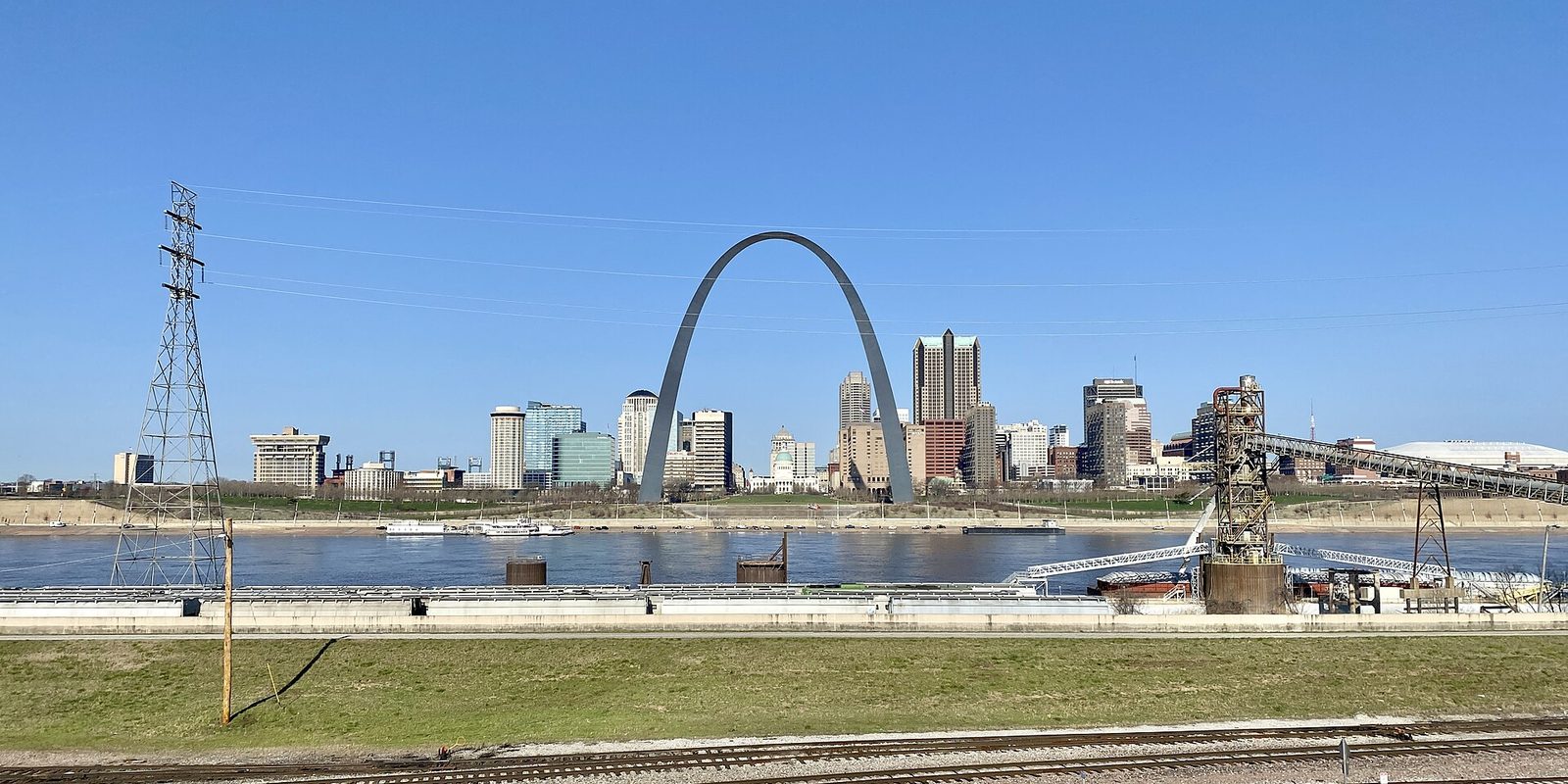 St. Louis skyline and the Gateway Arch across the Mississippi River under a cloudless blue sky, with railroad tracks and green grass in the foreground