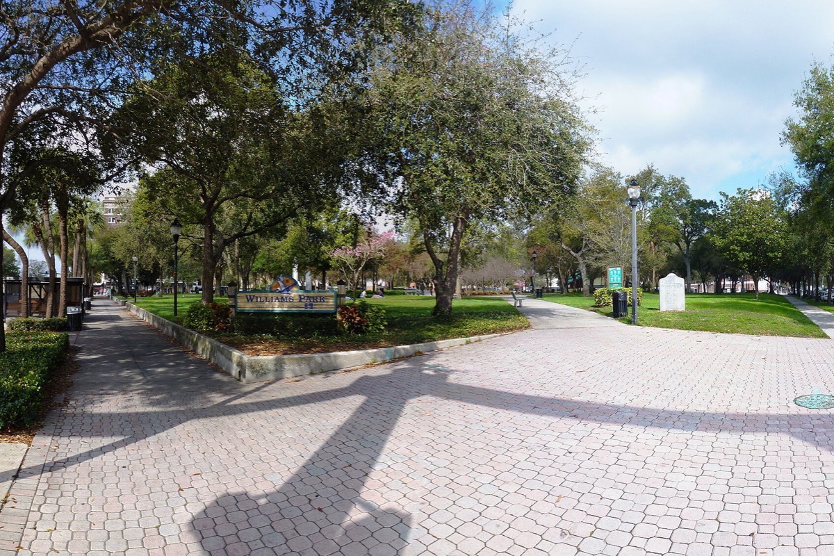 A broad paved path curving through a Florida park under mature shade trees, mown grass and low planting on both sides and buildings visible through the canopy.