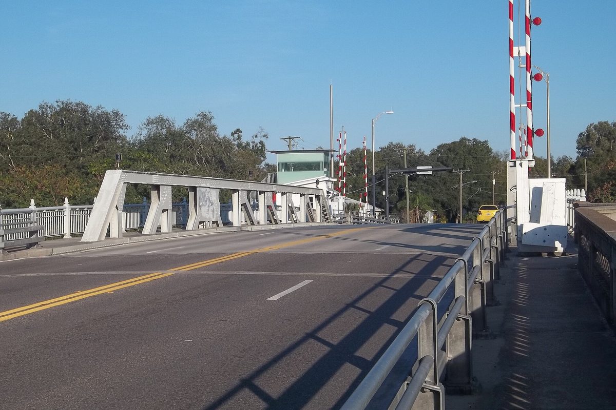 Tampa drawbridge roadway with concrete counterweight trusses, red-and-white striped warning gates and a glass operator house under a clear blue sky.