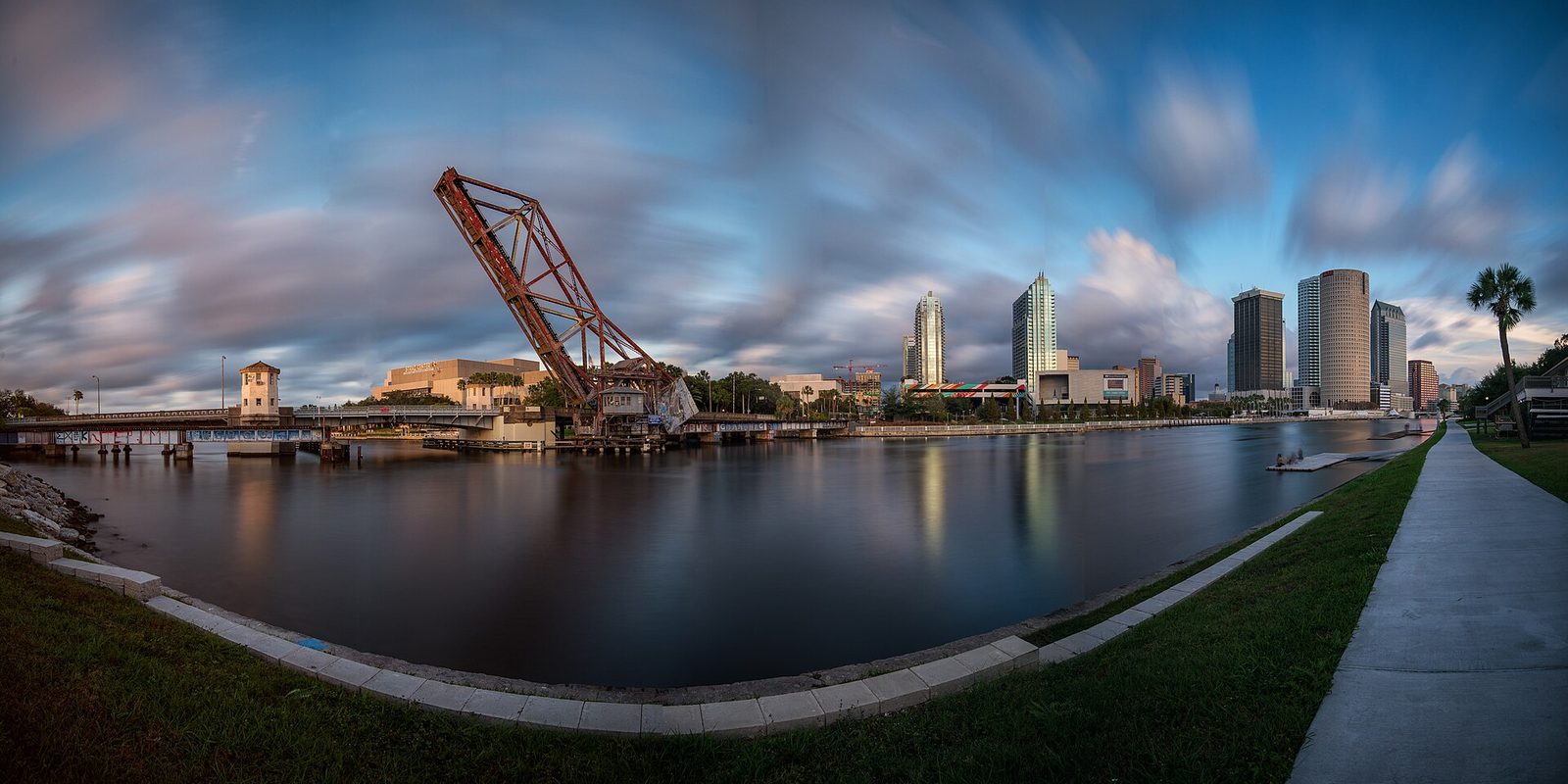 Tampa's downtown towers across the river at twilight, with a raised rust-red railroad lift bridge and streaked clouds above a riverside walkway
