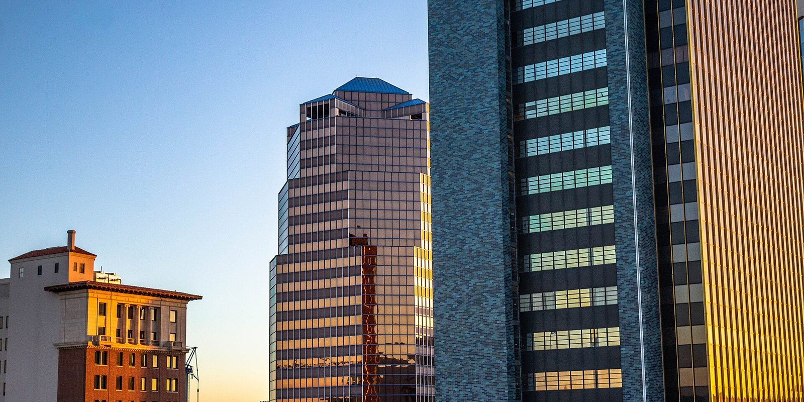 Tucson's downtown high-rises close up, glass and stone facades catching low golden sunlight beside a smaller older brick building under clear sky
