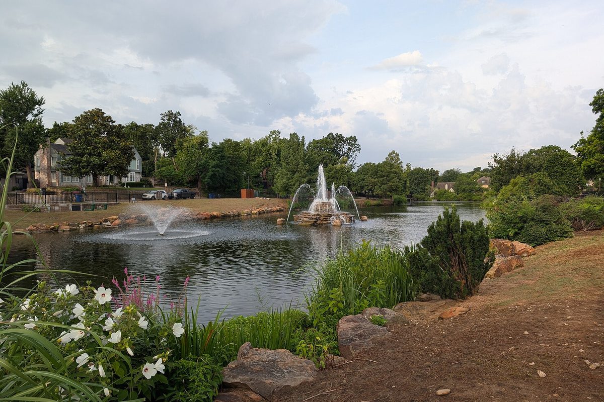 Tulsa park pond with two spraying fountains, boulder-lined banks, white hibiscus blooms in front and houses among leafy trees under a cloudy sky.