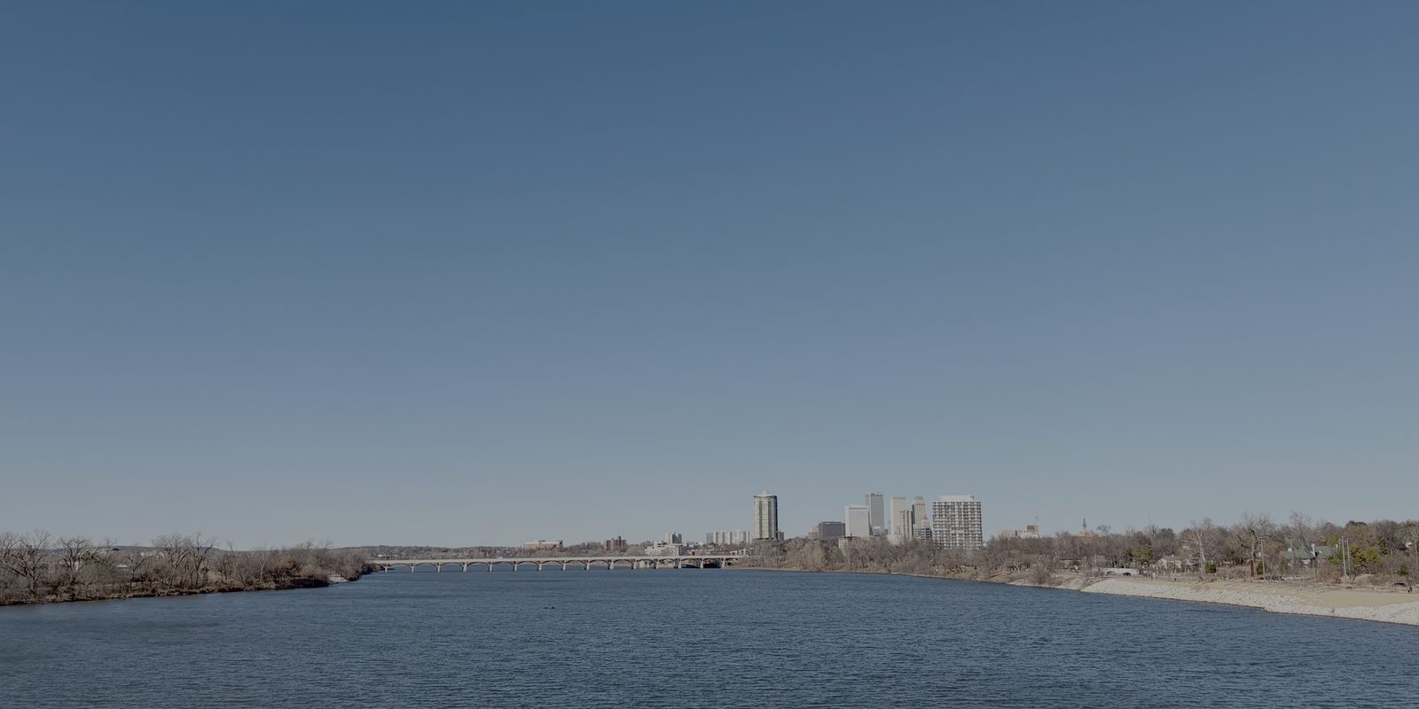 Tulsa seen across a wide river, downtown towers small on the horizon beyond a long low bridge, leafless trees lining both banks under a clear sky
