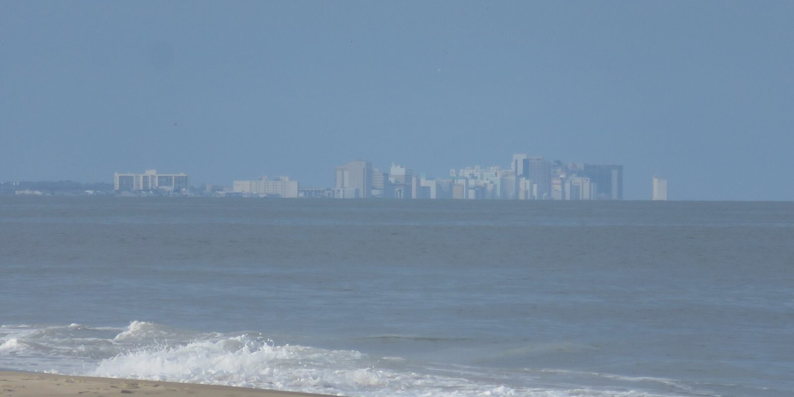 Virginia Beach's oceanfront hotel towers pale and hazy on the horizon across the water, with a wave breaking onto the sand in the foreground