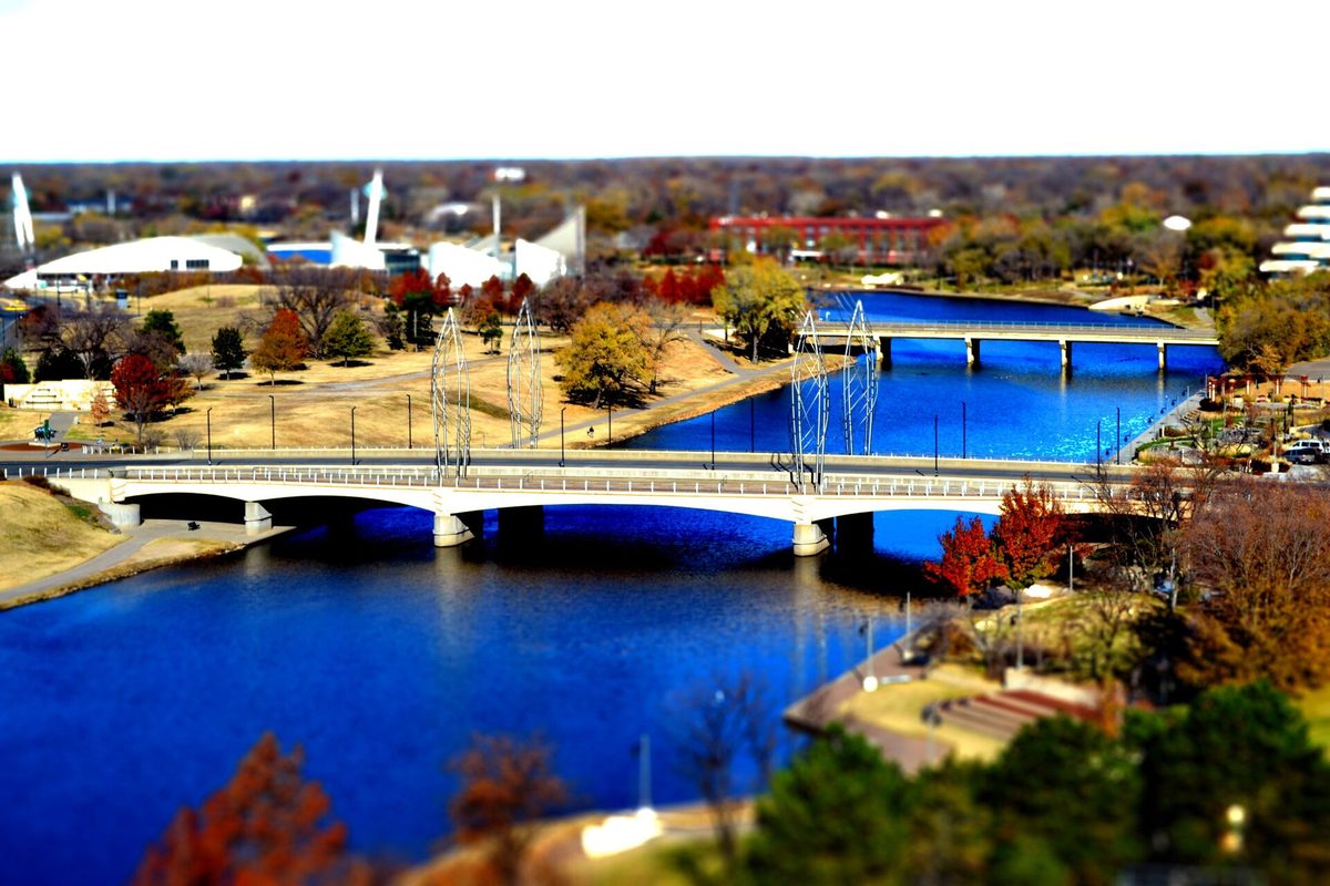 Wichita's Arkansas River curving past two low bridges with silver sculptures, autumn-brown parkland and a domed arena, in tilt-shift blur.