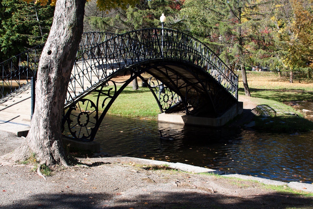 An ornate cast iron footbridge arching over a Massachusetts park pond, autumn colour in the trees behind it and the ironwork reflected in the still water.