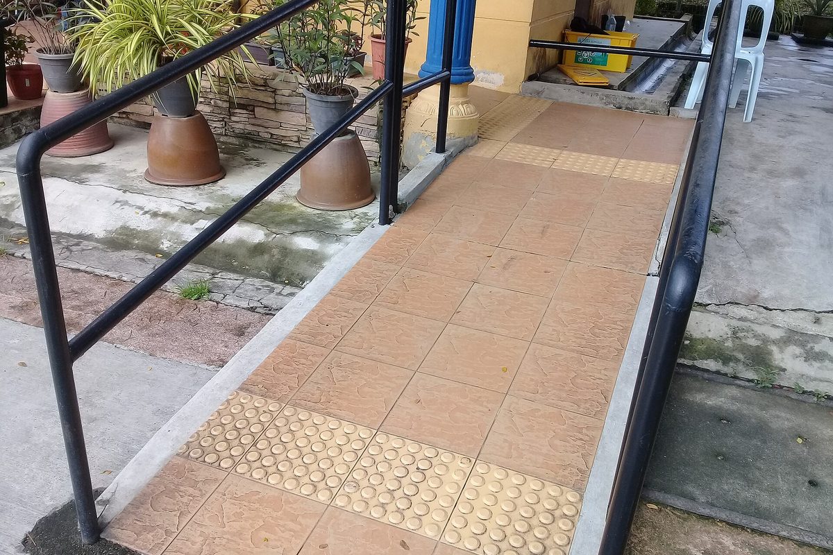 A tiled concrete ramp with black steel handrails and yellow raised-dot tactile paving, potted plants lining the wall beside it.