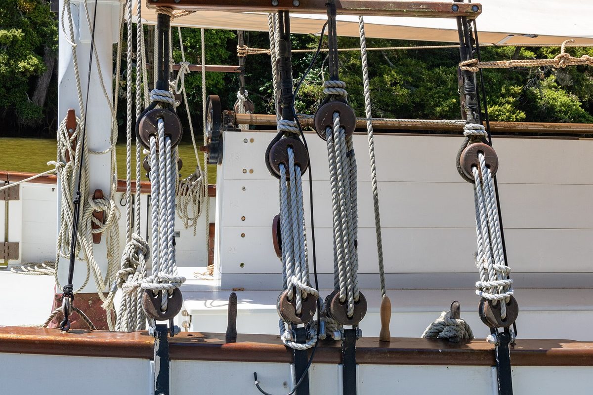 Coils of white rope running through weathered wooden pulley blocks in the rigging of a wooden sailboat, green trees behind.